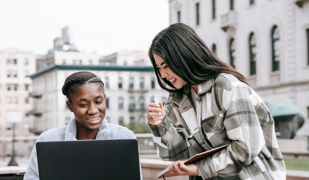 Twee jonge vrouwen kijken samen glimlachend naar een laptop buiten bij een universiteitsgebouw